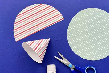 A dotted blue-printed circle of paper next to a stripey semi-circle, a stripey folded cone, scissors and glue stick.