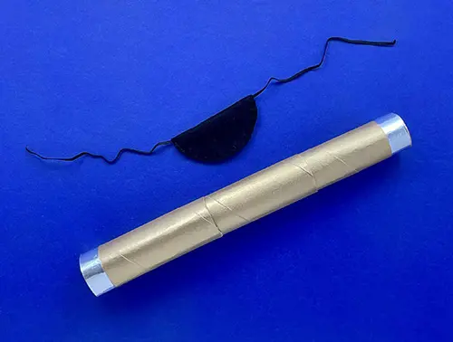 The gold DIY telescope is laid on a blue background below a black DIY fabric and elastic eye patch.
