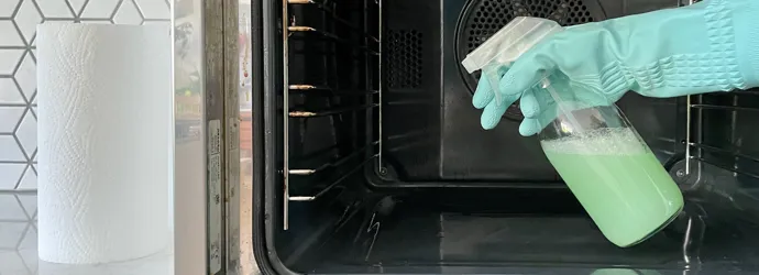 Young woman in apron cleaning the oven with spray and cloth