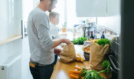 Man removing shopping from kitchen worktop with child by his side.