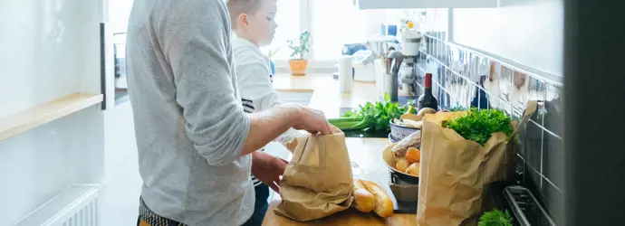 Man removing shopping from kitchen worktop with child by his side.