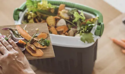 A woman's hand pushes vegetable peel into a small compost bin sitting on a countertop, demonstrating what you can put in a compost bin