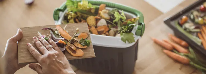 A woman's hand pushes vegetable peel into a small compost bin sitting on a countertop, demonstrating what you can put in a compost bin