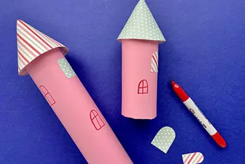 A red pen next to two pink cardboard towers, with paper windows stuck on and hand-drawn windows too.