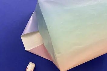 A rectangular cardboard box is being wrapped in pastel-coloured paper on a blue surface next to a stick of glue.