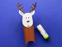 A brown painted cardboard tube has a paper reindeer head stuck on the top and is on a blue surface next to a glue stick.