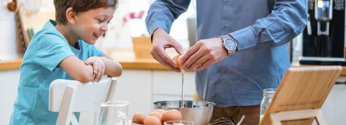 A man and a child joyfully crack eggs into a mixing bowl.