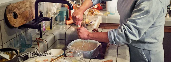 Woman with her sleeves rolled up washing dirty dishes in a full kitchen sink.
