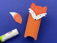 An orange painted paper tube has a fox head stuck on the top and is on a blue surface next to a glue stick and fox tail.