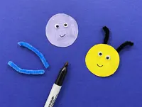 Circular blue and yellow paper circles have pipe cleaner antennae and googly eyes being attached with pen-drawn smiles.