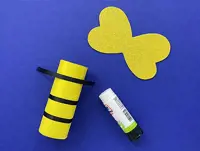 A yellow cardboard tube has black stripes being placed, next to a glue stick and yellow painted paper wings.