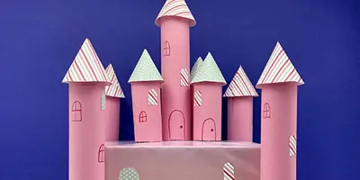 A pink cardboard castle with 7 kitchen towel tube towers around a rectangular base sits in front of a blue background.