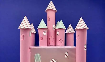 A pink cardboard castle with 7 kitchen towel tube towers around a rectangular base sits in front of a blue background.