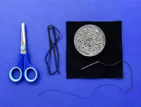 A square of black fabric, circular pin container, elastic, scissors, needle, and thread are laid out on a blue surface.