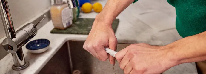 Hands of a person wearing green jumper cleaning the kitchen sink wringing out a wet kitchen towel.
