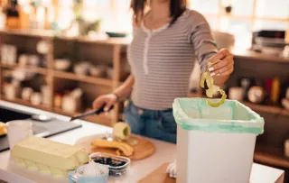 A woman chops vegetables and discards of the peels into a food waste bin.