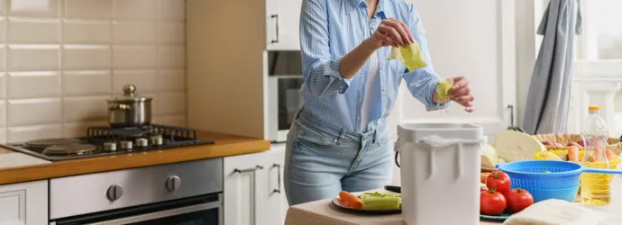 A woman in a blue shirt peels vegetables into a compost bin.