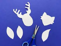 Paper reindeer and fox heads, owl wings and fox tail are laid on a blue surface with blue handled scissors.