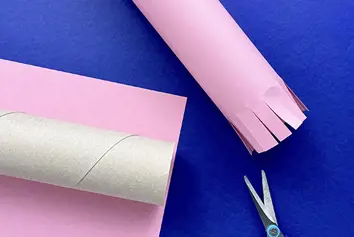 A cardboard tube on pink paper, next to a pair of scissors and pink-covered tube with a trimmed fringe on one end.