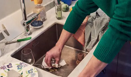 Man in a green jumper cleaning the kitchen sink with some tissue paper.
