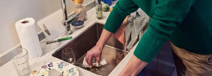 Man in a green jumper cleaning the kitchen sink with some tissue paper.