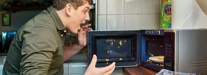Man looking at the food in the microwave after an explosion with a shocked expression.
