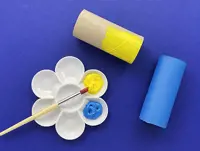 A container with blue and yellow paint and a paintbrush next to a blue cardboard tube and half painted yellow one.