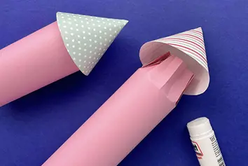 Two pink cardboard tubes are next to a glue stick and coloured paper cones ready to fit to the tubes as tower roofs.