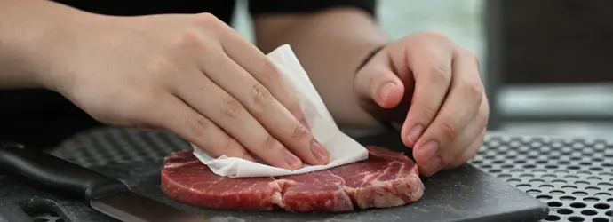 Person blotting a raw steak with a paper towel on a cutting board to remove moisture before cooking.