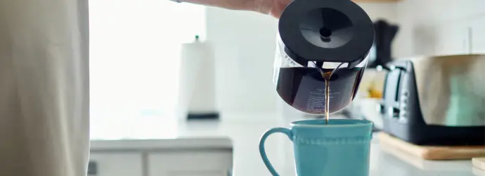 Person pours coffee from a glass carafe into a blue mug on a kitchen counter, with a toaster visible in the background.
