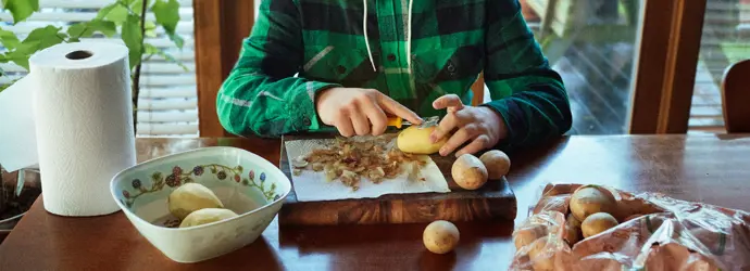Young boy peeling potatoes on a wooden table in kitchen.