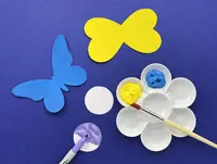 Paint is next to paper butterfly wings painted blue and bee wings in yellow. A pen is colouring a paper circle blue.