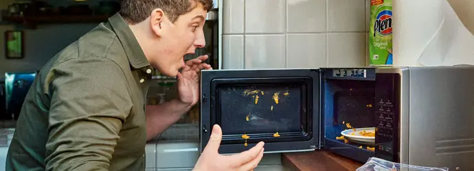 Ein Mann, der nach einer Explosion mit schockierter Miene auf das Essen in einer Mikrowelle schaut.
