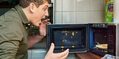 Ein Mann, der nach einer Explosion mit schockierter Miene auf das Essen in einer Mikrowelle schaut.
