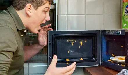 Ein Mann, der nach einer Explosion mit schockierter Miene auf das Essen in einer Mikrowelle schaut.
