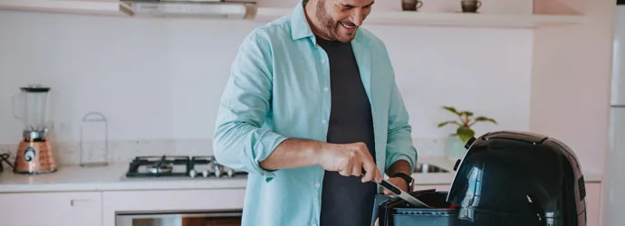 A smiling man stirs food in an airfryer, in a bright kitchen