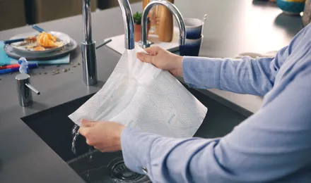 Man in a blue shirt holds a sheet of Plenty kitchen paper under a stainless still kitchen sink tap