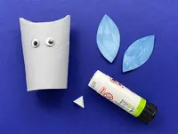 A grey painted owl paper tube has googly eyes on it with blue paper wings, a white beak and glue stick to the right.